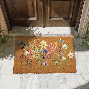 A colorful floral doormat with various flowers and butterflies placed on a tiled floor in front of a wooden door.