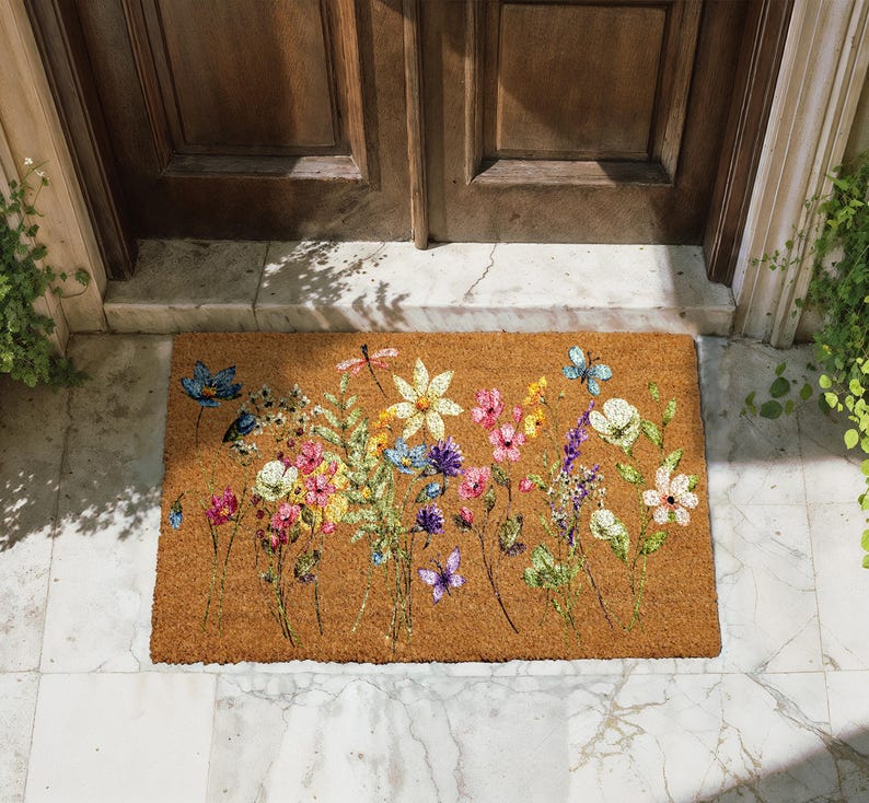 A colorful floral doormat with various flowers and butterflies placed on a tiled floor in front of a wooden door.