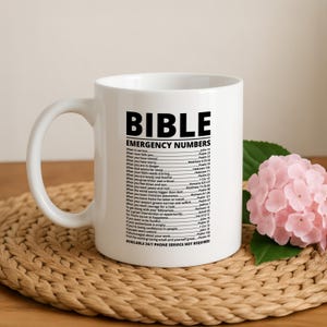 May include: White ceramic mug with a black handle featuring the text "BIBLE EMERGENCY NUMBERS" and a list of Bible verses for various situations. The mug is on a woven mat with a pink flower arrangement.