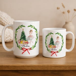 May include: White ceramic mugs with a Christmas-themed design. The design features a ballerina, Christmas tree, gifts, and a red bow. The name "Amelia" is written on the mug. One mug is larger than the other.