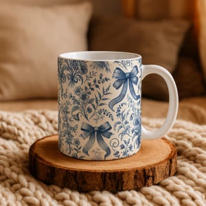 May include: A white ceramic mug featuring a blue toile-style pattern of intricate floral arrangements and decorative bows. The mug has a smooth cylindrical body with a curved handle on the right side, showcasing a detailed botanical print.