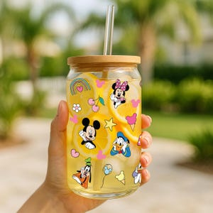 Peut inclure: Un récipient à boisson en verre transparent avec un couvercle en bois et une paille en verre. Le récipient est décoré de personnages de dessins animés colorés et rempli d'un liquide jaune et de tranches de citron.