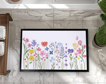 May include: A rectangular bath mat with a black border features a colorful floral design. The artwork showcases various flowers in shades of pink, yellow, blue, and purple, with a butterfly accent. The mat is placed on a marble tile floor.