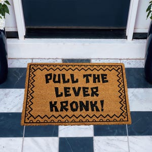 Puede incluir: Una alfombra rectangular hecha de fibra de coco natural. La alfombra presenta la frase "PULL THE LEVER KRONK!" en letras negras en negrita. El borde está decorado con un patrón de zigzag negro. La alfombra está colocada sobre un suelo embaldosado.