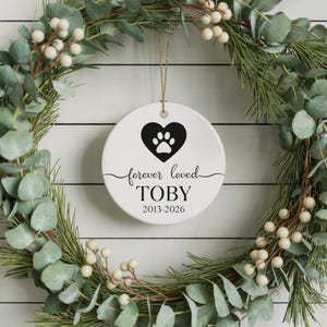 Può includere: Ornamento rotondo in ceramica bianca con un cuore nero e un'impronta di zampa. Le parole "forever loved" e il nome "TOBY" sono stampati in nero. Sospeso da un filo d'oro, è circondato da una ghirlanda verde.