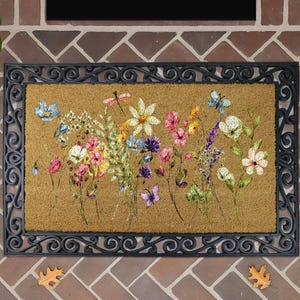 A decorative doormat with a floral design, featuring various colorful flowers and plants. The doormat is placed on a brick wall, and there are some leaves on the ground around it.