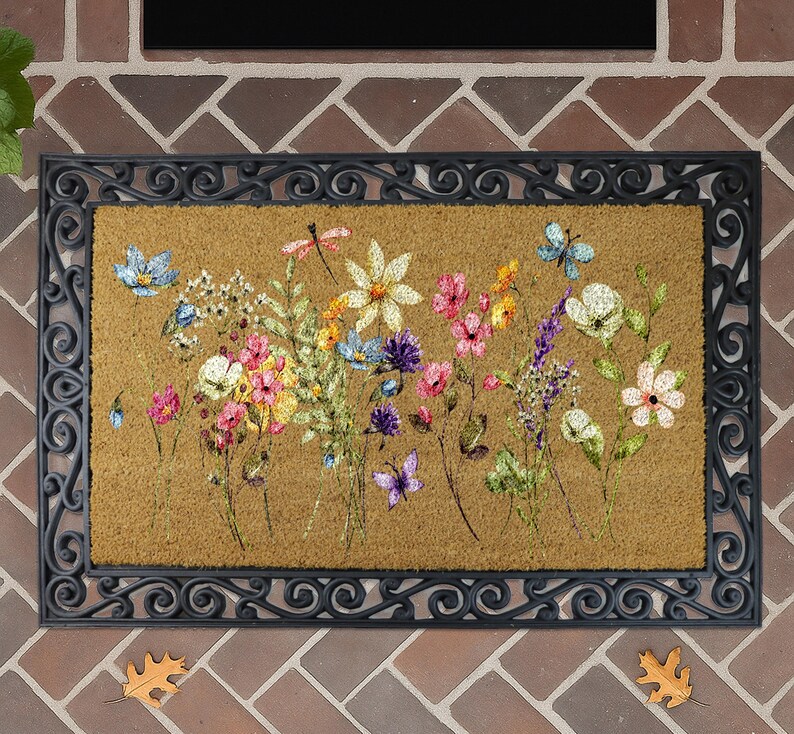 A decorative doormat with a floral design, featuring various colorful flowers and plants. The doormat is placed on a brick wall, and there are some leaves on the ground around it.