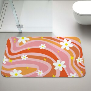 May include: A rectangular bath mat with a retro design. The mat features a wavy pattern in shades of orange, pink, and white, with white daisy flowers and yellow star accents. The mat is placed in a bathroom setting.