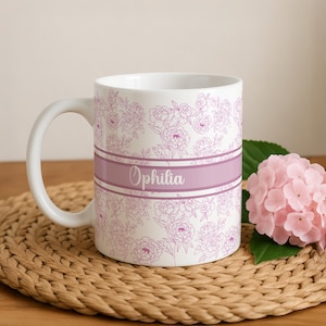 May include: A white ceramic mug featuring a repeating pattern of delicate purple floral line drawings. A horizontal band in a solid light purple shade wraps around the center, displaying the name Ophilia in a white cursive script font.