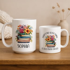 May include: Two white ceramic mugs with floral and book designs. The larger mug has the name "Sophia" and the smaller mug has the text "There's no such thing as too many books". Both mugs feature colorful flower arrangements.