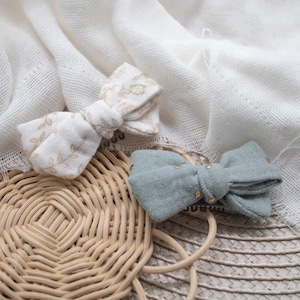 Puede incluir: Dos lazos de pelo de tela, uno con un patrón floral en beige y blanco, y el otro en azul claro sólido con detalles de purpurina dorada. Los lazos están sobre una superficie de mimbre tejido.