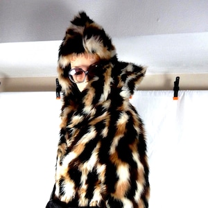 May include: A long, hooded coat with a faux fur leopard print design in black, brown, and cream. The coat features a black belt and a matching black bag. The person is wearing sunglasses.