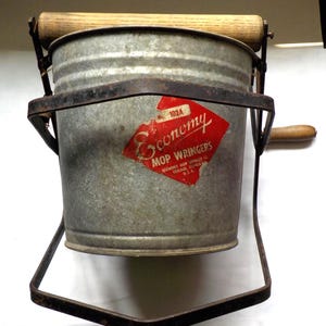 May include: Vintage metal mop wringer with a galvanized bucket and wooden handles. A red and white label reads "Economy Mop Wringers, Chicago, U.S.A." The wringer has a metal frame and is designed to squeeze water from a mop.