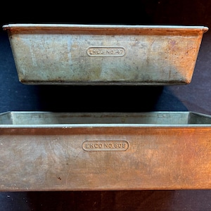 May include: Two vintage rectangular metal loaf pans. The top pan is marked "EKCO NO. 47", and the bottom pan is marked "EKCO NO. 608". The pans have a worn, aged appearance, with a metallic silver color and signs of use.