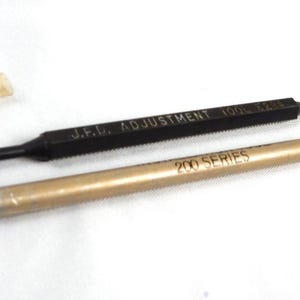 May include: A black J.F.D. ADJUSTMENT 100L 6284 tool with a small translucent cap and a gold-colored 200 SERIES tool with a silver tip. Both tools are slender, cylindrical, and feature printed text along their lengths on a plain white surface.