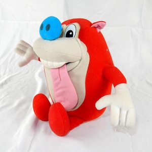 May include: A plush toy of a cartoon character with a red body, a light beige face, and a blue nose. The character has a pink tongue sticking out and white gloves. The toy is sitting on a white surface.