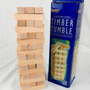May include: A wooden Timber Tumble game with a tower of rectangular blocks next to a blue metal container. The container has the game title and a graphic of the game. The game is for ages 8+ and 2+ players.