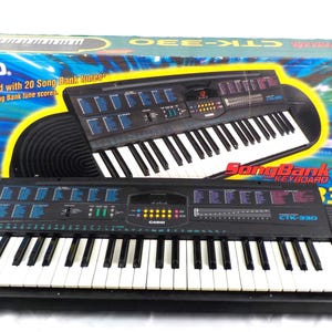 May include: A black Casio CTK-330 SongBank keyboard with white keys and blue control buttons. The keyboard is on a black base. The box behind it shows a similar keyboard and lists features like 49 full-size keys and 100 tones.