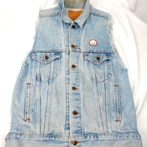May include: Light blue denim vest with a button-up front, two chest pockets, and two side pockets. The vest has a distressed look with frayed edges and a small smiley face patch. The vest has a collar and a brown label.