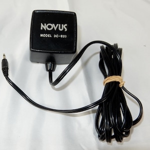 May include: Black Novus Model DC-920 power adapter with a black cord and a 3.5mm jack. The cord is bundled with a tan twist tie. The adapter has the brand name "NOVUS" printed in white.