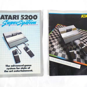 May include: Two vintage Atari 5200 SuperSystem game console brochures. One features the console and controllers on a light blue background with the text "ATARI 5200 SuperSystem". The other brochure has the console and controllers on a black and white checkered background.