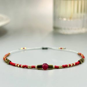 Può includere: Un braccialetto di perline rosse con dettagli dorati e una perlina rossa centrale sfaccettata. Il braccialetto è infilato su un cordino bianco e presenta perline quadrate color oro. Un gioiello delicato.