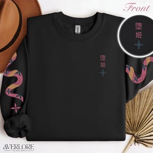 Könnte beinhalten: Schwarzes Sweatshirt mit einem Schlangenmuster auf den Ärmeln und japanischem Text und floralem Design auf der Vorderseite. Die Ärmel haben ein rosa, orangefarbenes und gelbes Schlangenmuster. Der Text auf der Vorderseite ist rosa und das florale Design ist blau.