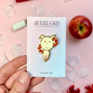 Puede incluir: Un pin de esmalte dorado con un personaje de dibujos animados con cuerpo crema y detalles de flores rojas. El pin está sobre una tarjeta blanca con el texto "AVERLORE FAN MADE MERCH" y "ENAMEL PIN". El fondo es rosa con una manzana y pétalos de flores.