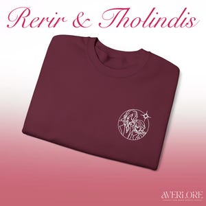Puede incluir: Una camiseta granate con el texto "Rerir & Tholindis" en una fuente decorativa. La camiseta presenta un gráfico circular blanco de dos figuras y una estrella. La esquina inferior derecha tiene el texto "AVERLORE FAN MADE MERCH".