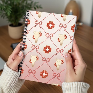 Puede incluir: Cuaderno con espiral con un patrón repetido de caras de cerdos de dibujos animados, lazos rojos y detalles florales sobre un fondo rosa y crema degradado. El cuaderno está abierto, mostrando el diseño.