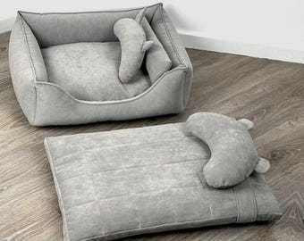 Puede incluir: Juego de dos camas para mascotas en gris claro. El juego incluye una cama rectangular con laterales elevados y una almohada a juego. También se incluye una cama rectangular plana separada con una almohada curva. Las camas están hechas de un material suave y afelpado.