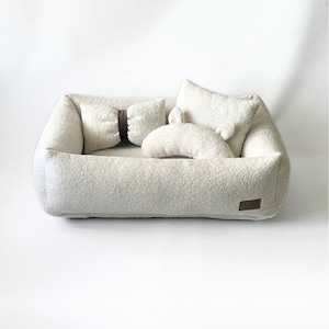 May include: A cream-colored pet bed with a rectangular shape and soft, textured fabric. It includes several matching pillows and a small, brown leather tag. The bed is designed for comfort and relaxation for small pets.