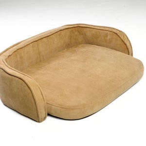 Puede incluir: Una cama para mascotas de color beige con respaldo y laterales curvos. La cama tiene un aspecto suave y afelpado y está diseñada para la comodidad. El color neutro la hace adecuada para varios estilos de decoración.