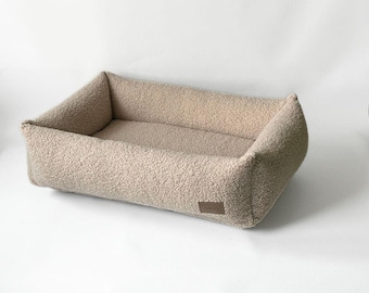 Puede incluir: Una cama rectangular para mascotas hecha de tela de forro polar sherpa beige texturizada. La cama cuenta con lados elevados para soporte y una pequeña etiqueta de marca rectangular marrón cosida en la esquina inferior frontal.