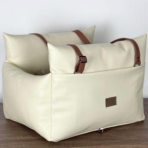 May include: Cream-coloured pet car seat with brown leather straps and accents. The seat has a rectangular shape with a raised back and sides, and a small brown label is attached to the front. Designed to keep pets safe and secure while travelling.