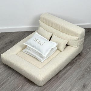 May include: Cream-colored pet bed with a rectangular base and a raised backrest. Includes a stack of white pillows and a pillow with the name "Maxi" embroidered on it. The bed is made of a soft, textured material.