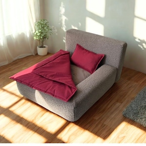 May include: A small, grey pet bed with a burgundy blanket and pillow. The bed has a low profile and a rounded back. The blanket is folded over the bed, and the pillow is placed on the bed. The bed is on a wooden floor.