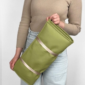 May include: A green clutch with two beige straps. The clutch is rectangular and folded over. The person in the image is wearing a tan turtleneck sweater and light blue jeans.