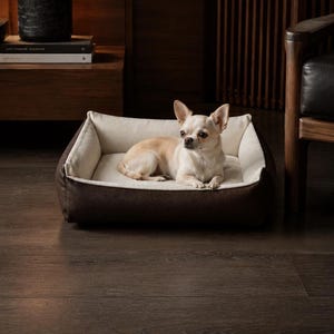以下が含まれることがあります： ダークブラウンの質感のある外側とクリーム色の内側クッションを備えた長方形のペット用ベッド。大きな耳と鋭い目を持つ小さなライトタンカラーのチワワが、ベッドの中央で右側を向いて休んでいます。