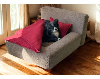 Op de afbeelding: Een kleine, langharige zwart-witte hond rust in een rechthoekig huisdierbed. Het bed is bekleed met een gestructureerde, lichtbruine, krullende stof en is voorzien van een felrood, rechthoekig kussen over het onderlichaam van de hond.