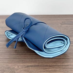 Op de afbeelding: Een opgerolde, gewatteerde, marineblauwe leren make-up kwastrol met een lichtblauwe binnenkant. De rol is vastgezet met een bijpassende marineblauwe stropdas. De rol ligt op een houten oppervlak.