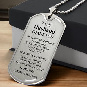 May include: A silver dog tag pendant with a ball chain necklace. The tag has the words "To My Husband THANK YOU" and a loving message engraved on it. The tag is displayed on a wooden surface next to a black box.