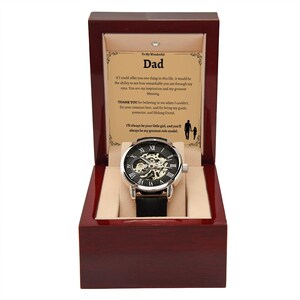 May include: A mechanical watch with a black leather band is displayed in a wooden box. The watch has a silver-colored case and a black face with visible gears. The box contains a card with text that reads "To My Wonderful Dad."