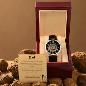 May include: A black leather strap watch with a silver case and a skeleton dial is displayed inside a red wooden box. A card with text is placed in front of the box. The watch has Roman numerals.