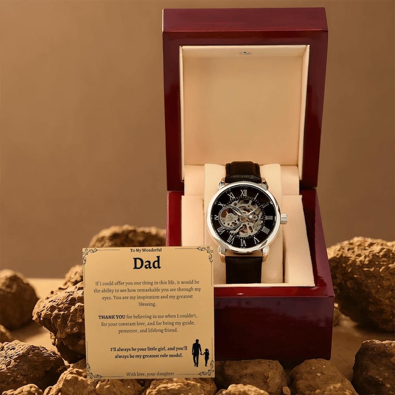 May include: A black leather strap watch with a silver case and a skeleton dial is displayed inside a red wooden box. A card with text is placed in front of the box. The watch has Roman numerals.
