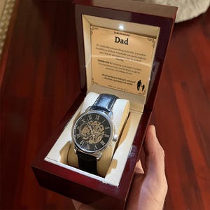 May include: A wristwatch with a black leather band and a silver-tone case is displayed inside a wooden box. The watch face has Roman numerals and a visible mechanical movement. The box contains a printed message that reads "To My Wonderful Dad."