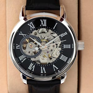May include: A silver-toned, skeleton-dial wristwatch with a black face and Roman numeral hour markers. The watch features visible gears and a black leather band. The watch is a classic design.