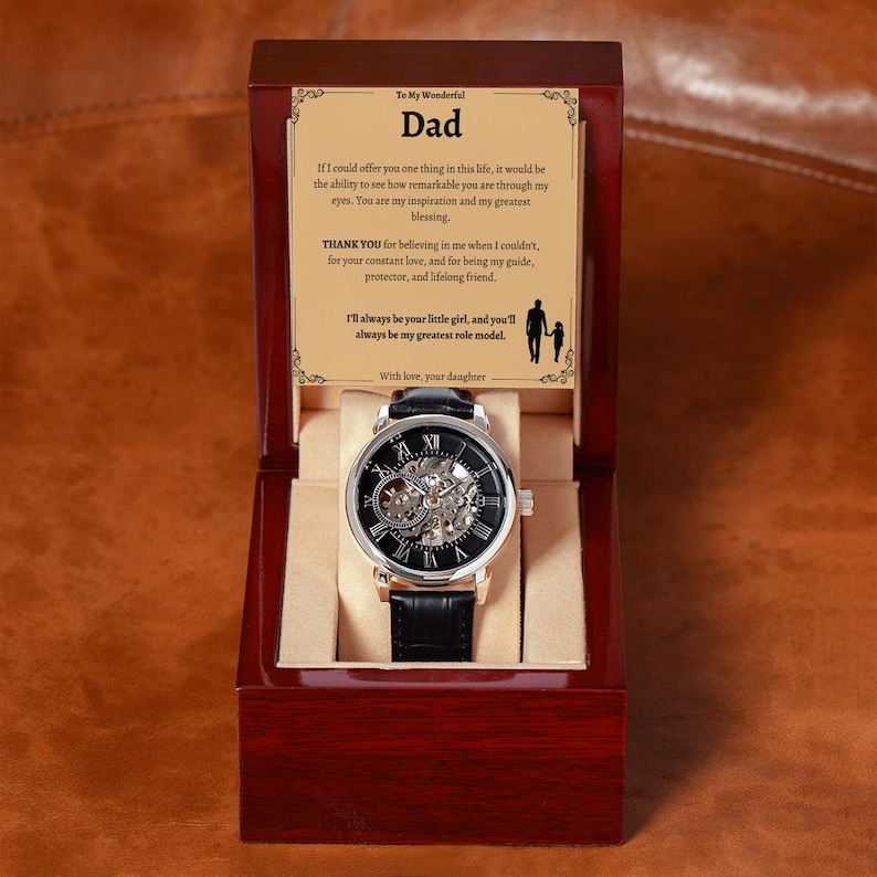 May include: A mechanical watch with a black leather band is displayed inside a wooden box. The watch has a silver-colored case and a black face with visible gears. The box contains a message to "Dad" from "your daughter".