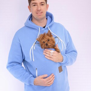 May include: A pale blue hoodie with a kangaroo pocket, a brown patch, and a white zip. A small, brown dog is in the pocket. The hoodie is part of a matching set with pale blue tracksuit bottoms.