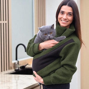 May include: A green and black pet carrier jacket holds a grey cat. The jacket has a high collar and a front pocket designed to carry a cat. The person holding the jacket is smiling.